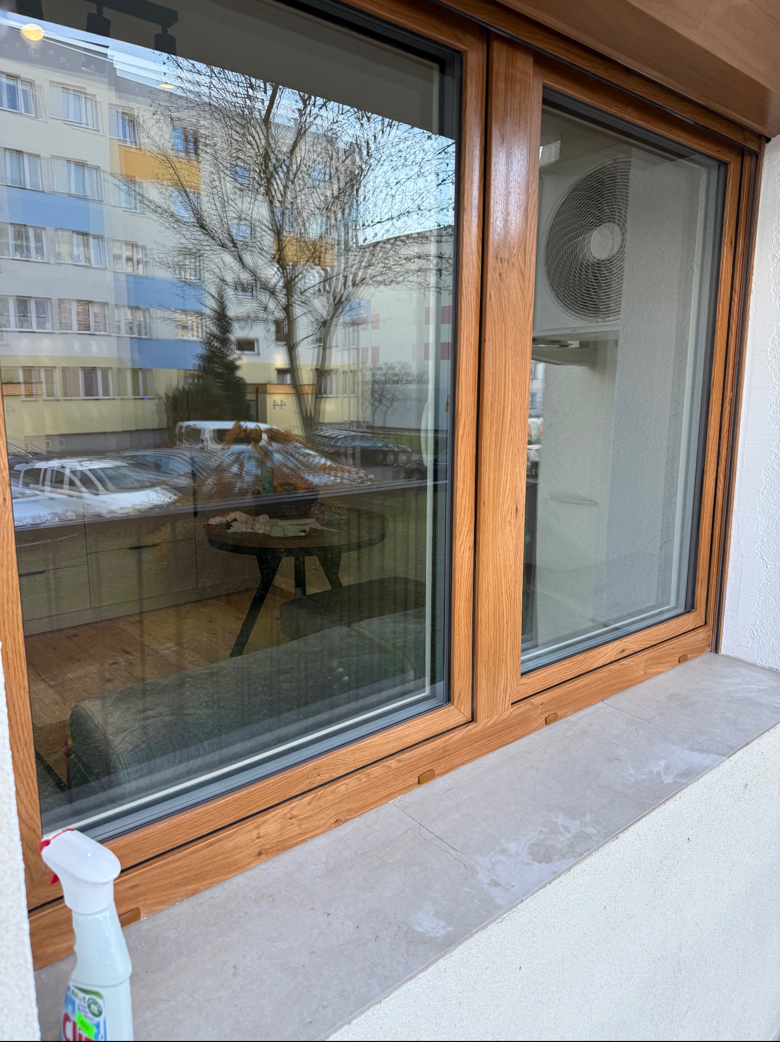 Drewniane okno z widokiem na blok i drzewo, na parapecie butelka ze środkiem czyszczącym. Widoczny klimatyzator na zewnątrz. Ujęcie z dołu, podkreślające detale ramy okiennej.