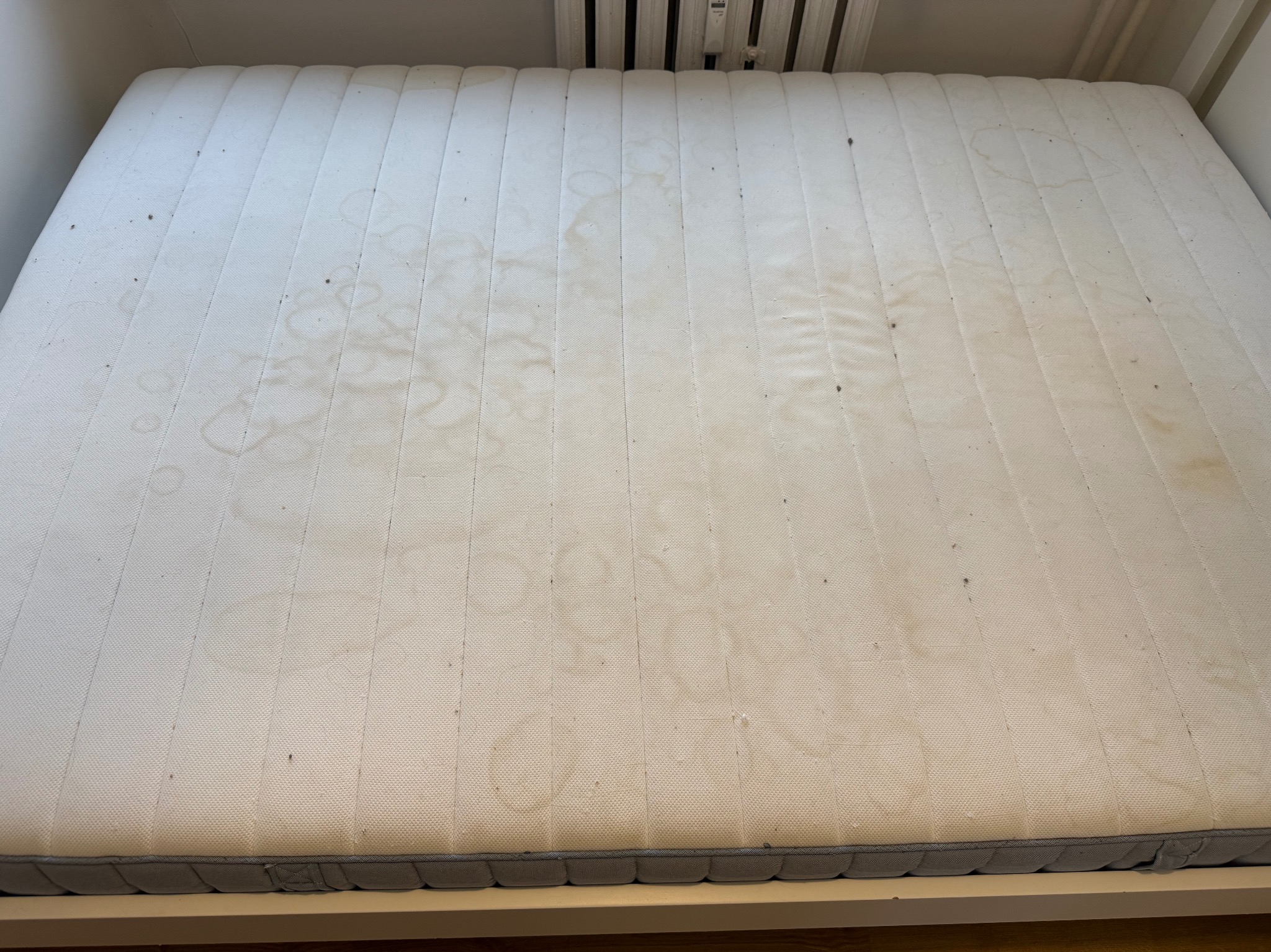 Zabrudzony materac z widocznymi plamami i drobnymi zabrudzeniami, umieszczony w ramie łóżka. Widoczne pionowe przeszycia i szara lamówka.
