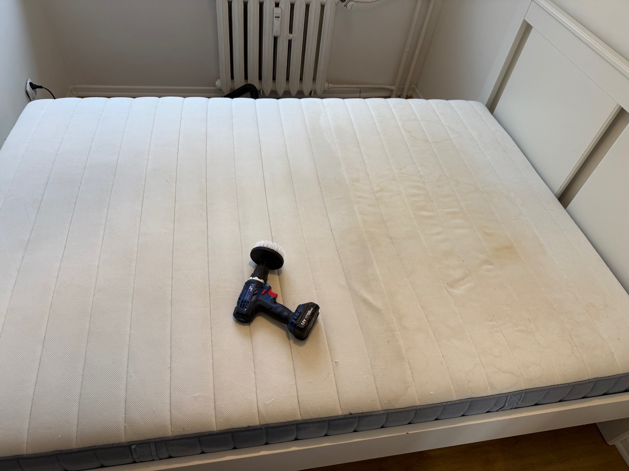 Materac z plamami czyszczony w Wolsztynie. Na materacu leży wiertarka z okrągłą szczotką do czyszczenia tapicerki. Widoczne ślady po czyszczeniu.