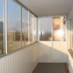 Przeszklony balkon z białymi ramami, widok na budynek mieszkalny w oddali, pionowe panele ścienne z tworzywa sztucznego.