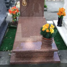 Granitowy nagrobek w odcieniach brązu z płaskorzeźbą twarzy i kwiatami w wazonach, ustawiony na cmentarzu.