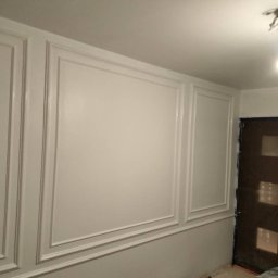 Tomasz Szaniaw - Biała ściana z dekoracyjnymi panelami i listwami, oświetlona lampą sufitową. Drzwi zabezpieczone folią malarską. Detal wykończenia wnętrza.