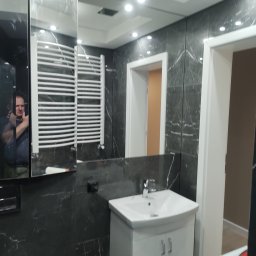 Eko-Tech Budownictwo Premium - Elegancka łazienka z czarnymi płytkami imitującymi marmur, białą szafką z umywalką i grzejnikiem drabinkowym. W lustrze odbija się osoba robiąca zdjęcie.