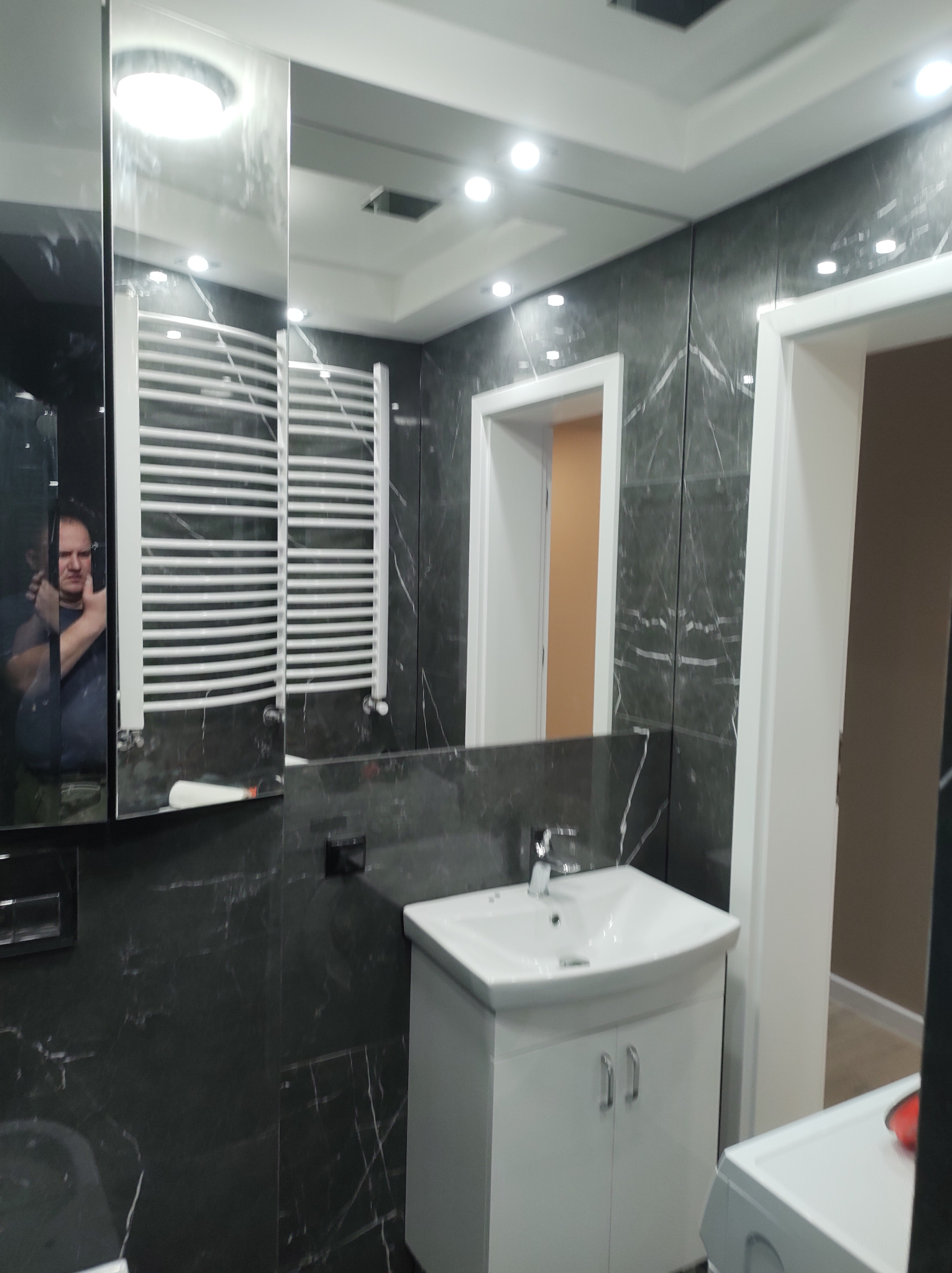 Elegancka łazienka z czarnymi płytkami imitującymi marmur, białą szafką z umywalką i grzejnikiem drabinkowym. W lustrze odbija się osoba robiąca zdjęcie.