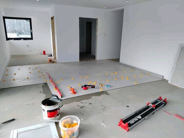 Układanie płytek w jasnym wnętrzu. Widoczny fragment ułożonej podłogi, narzędzia glazurnicze: poziomica, przyssawki, wiadro z klejem i gąbką, oraz gilotyna do cięcia płytek.