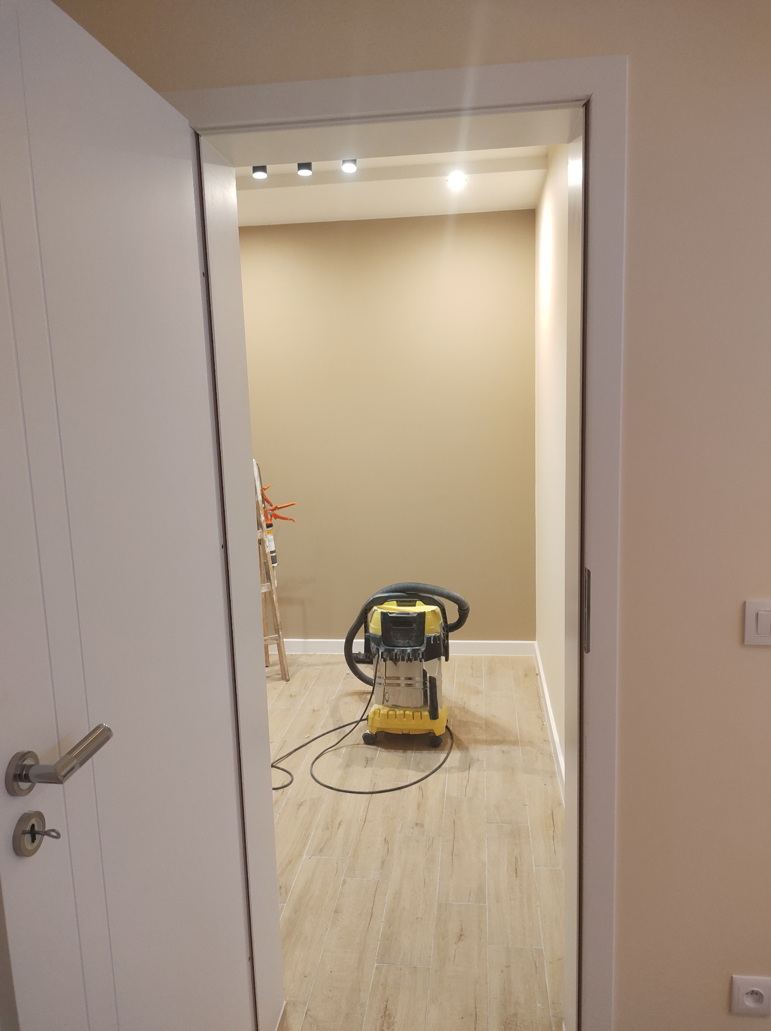 Wnętrze po remoncie: beżowe ściany, drewnopodobna podłoga, białe listwy przypodłogowe. Widok zza otwartych drzwi, w tle drabina i odkurzacz przemysłowy.