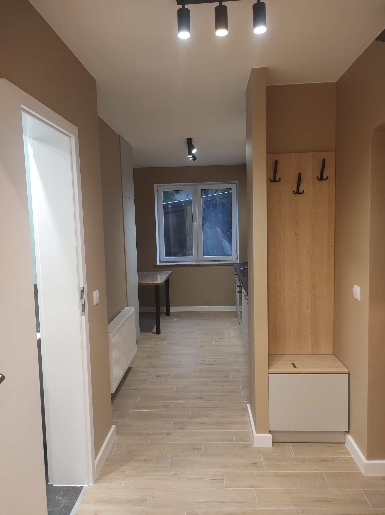 Nowoczesny przedpokój w beżach: drewniany panel z wieszakami, biała szafka, ceramiczna podłoga imitująca drewno, oświetlenie punktowe i widok na okno.