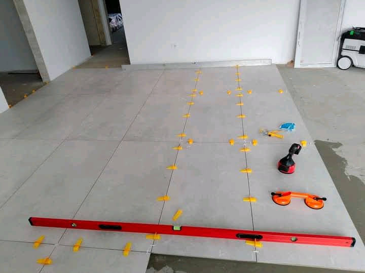 Układanie jasnych płytek podłogowych z użyciem poziomicy i klipsów w Gorzowie Wielkopolskim. Widoczne narzędzia glazurnicze i fragment odkurzacza przemysłowego.