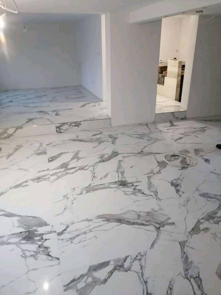 Biała marmurowa podłoga z szarymi żyłami w nowym, jasnym wnętrzu. Widoczne przejście do kolejnego pomieszczenia i fragment zwisającego kabla na podłodze.