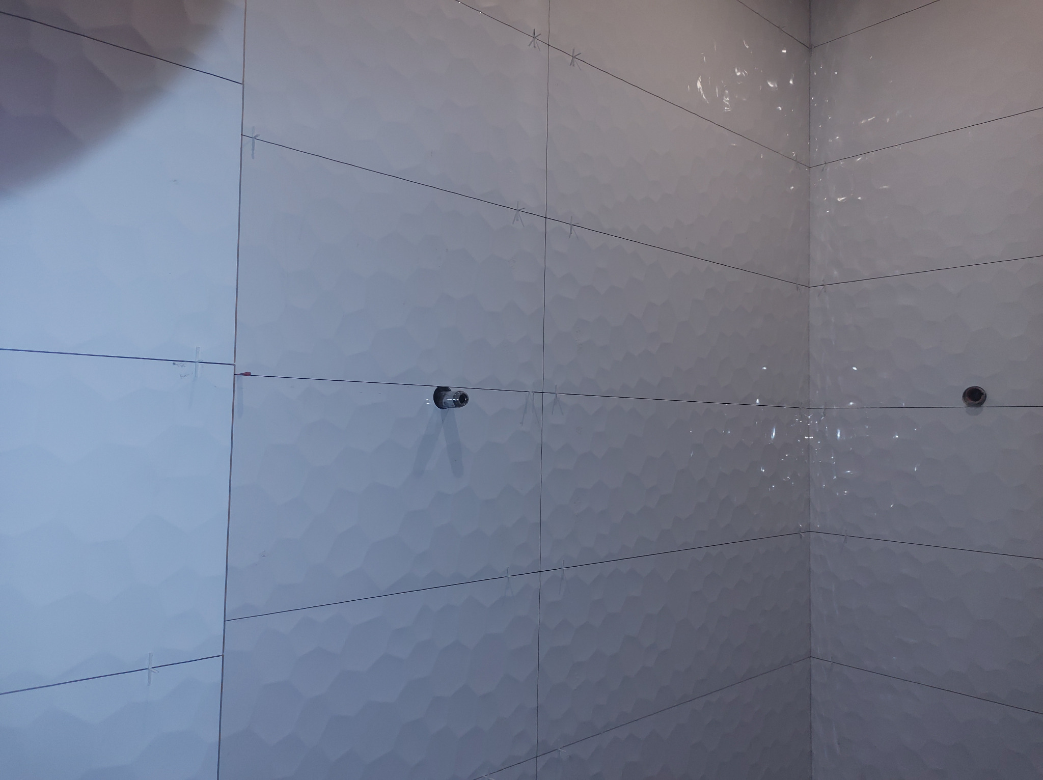 Ściana wyłożona białymi, trójwymiarowymi płytkami w łazience. Widoczne krzyżyki dystansowe i otwory na armaturę. Układ poziomy, fuga w kolorze szarym.