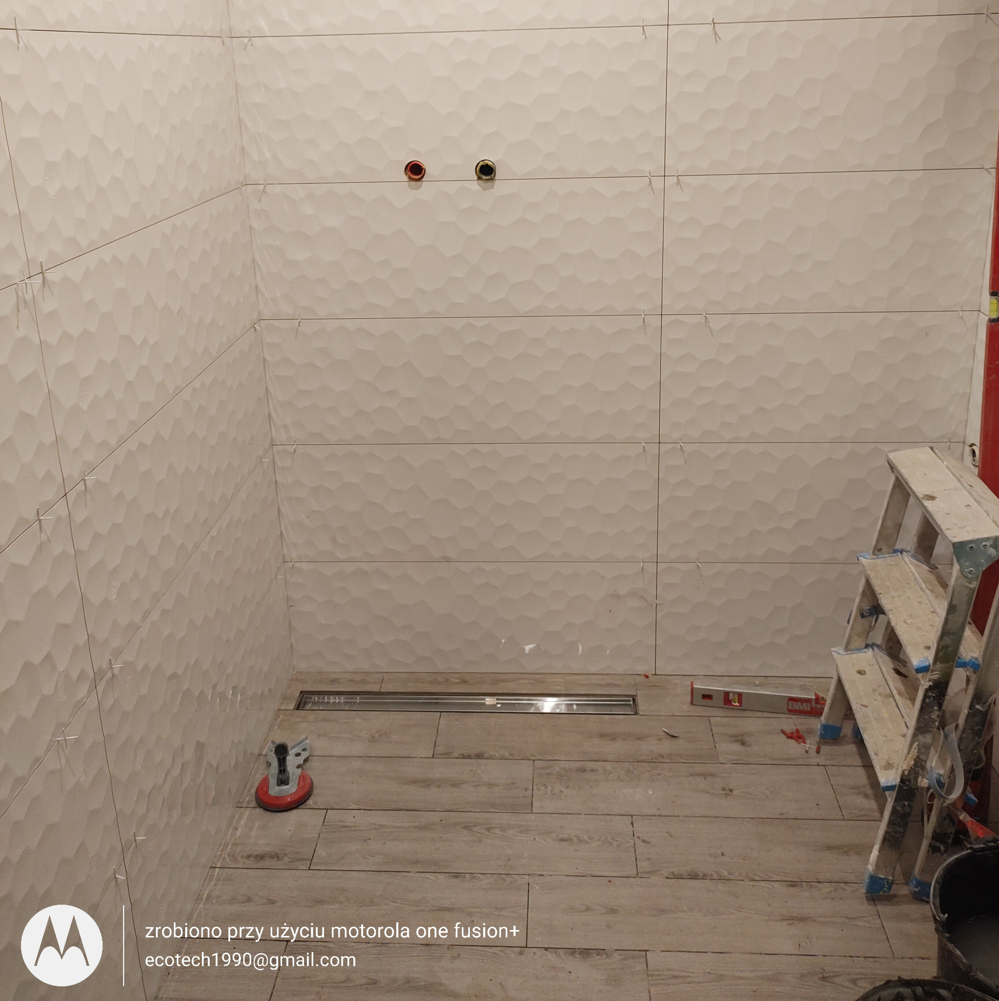 Układanie beżowych, strukturalnych płytek w łazience. Widoczny odpływ liniowy, poziomica i drabina. Praca w toku, płytki ułożone do połowy wysokości ścian.