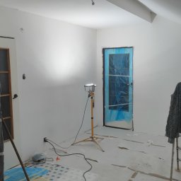 Tomasz Wieliczko - Białe ściany w pokoju po malowaniu, zabezpieczone drzwi folią, lampa budowlana na statywie, częściowo odsłonięta podłoga z parkietem. Widok na wnętrze w trakcie prac.