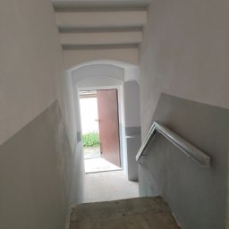 Tomasz Wieliczko - Klatka schodowa po remoncie w Lwówku Śląskim. Ściany pomalowane na szaro i biało, widoczne schody i fragment poręczy. Otwarta, brązowa  furtka na końcu schodów.
