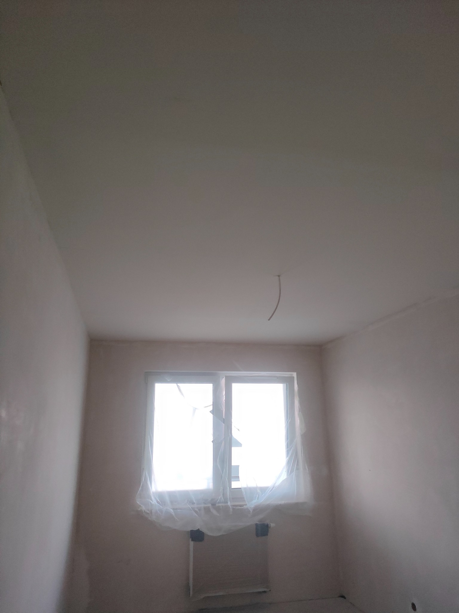 Pokój w trakcie prac wykończeniowych: ściany przygotowane do malowania, okno zabezpieczone folią, widoczne ślady szpachlowania. Minimalistyczne ujęcie wnętrza.