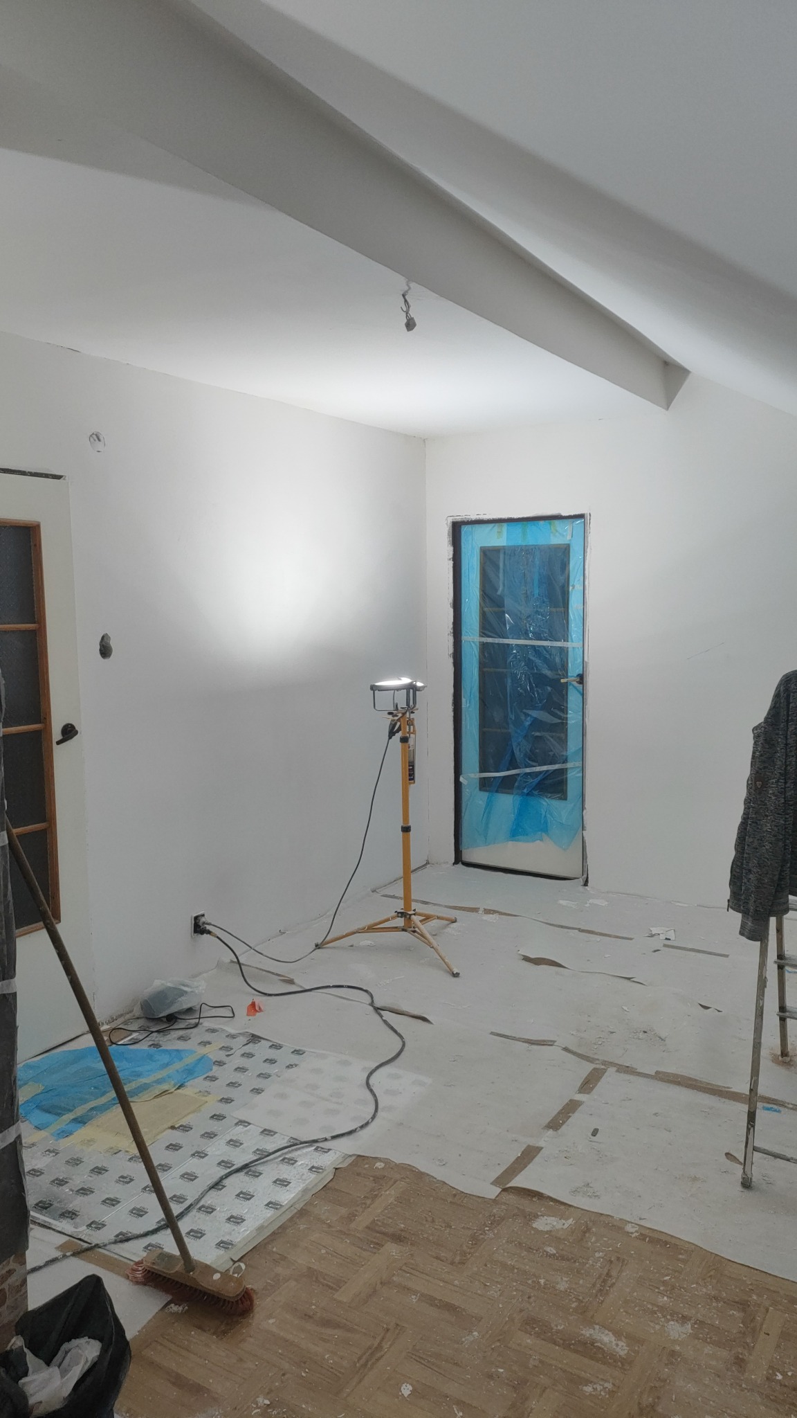 Białe ściany w pokoju po malowaniu, zabezpieczone drzwi folią, lampa budowlana na statywie, częściowo odsłonięta podłoga z parkietem. Widok na wnętrze w trakcie prac.