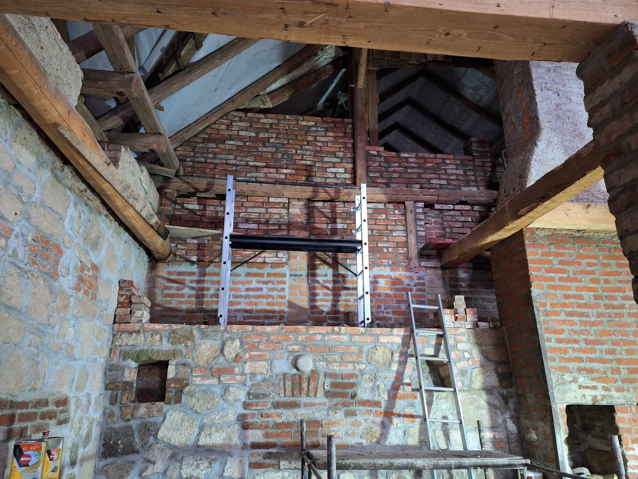Wnętrze w trakcie renowacji: surowe mury z cegły i kamienia, drewniane belki stropowe, rusztowania. Widoczne ślady prac remontowych, surowy stan wykończenia.