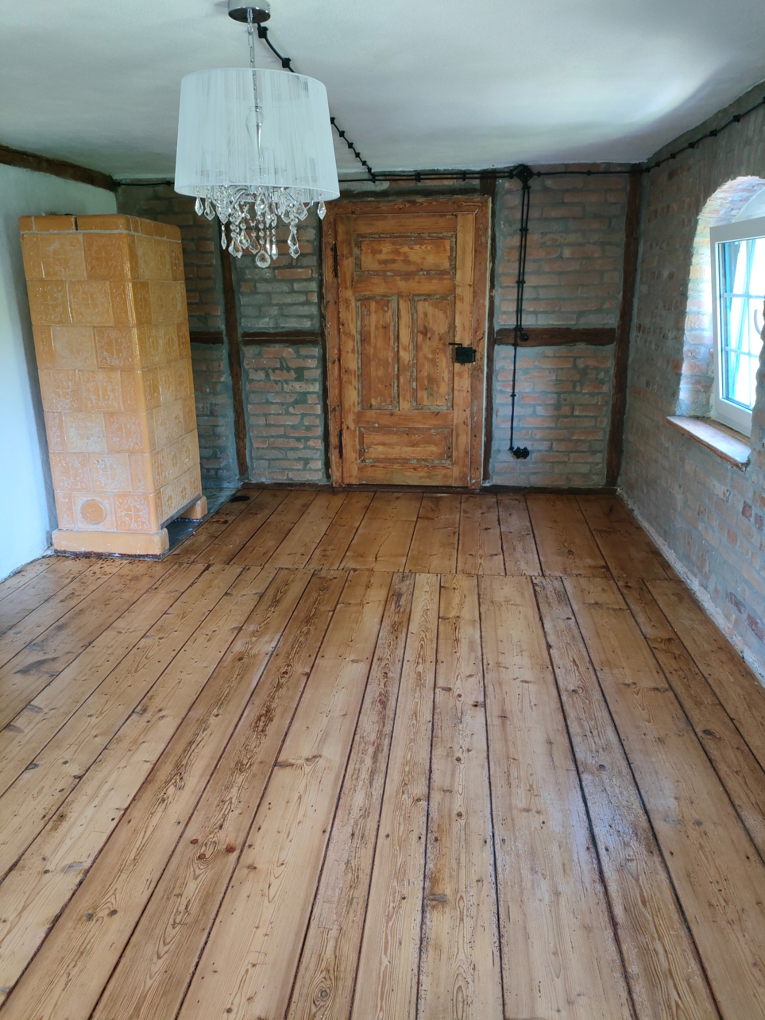 Pokój po renowacji: drewniana podłoga, ceglane ściany z drewnianymi belkami, piec kaflowy i ozdobny żyrandol. Widoczne ślady użytkowania dodają charakteru.