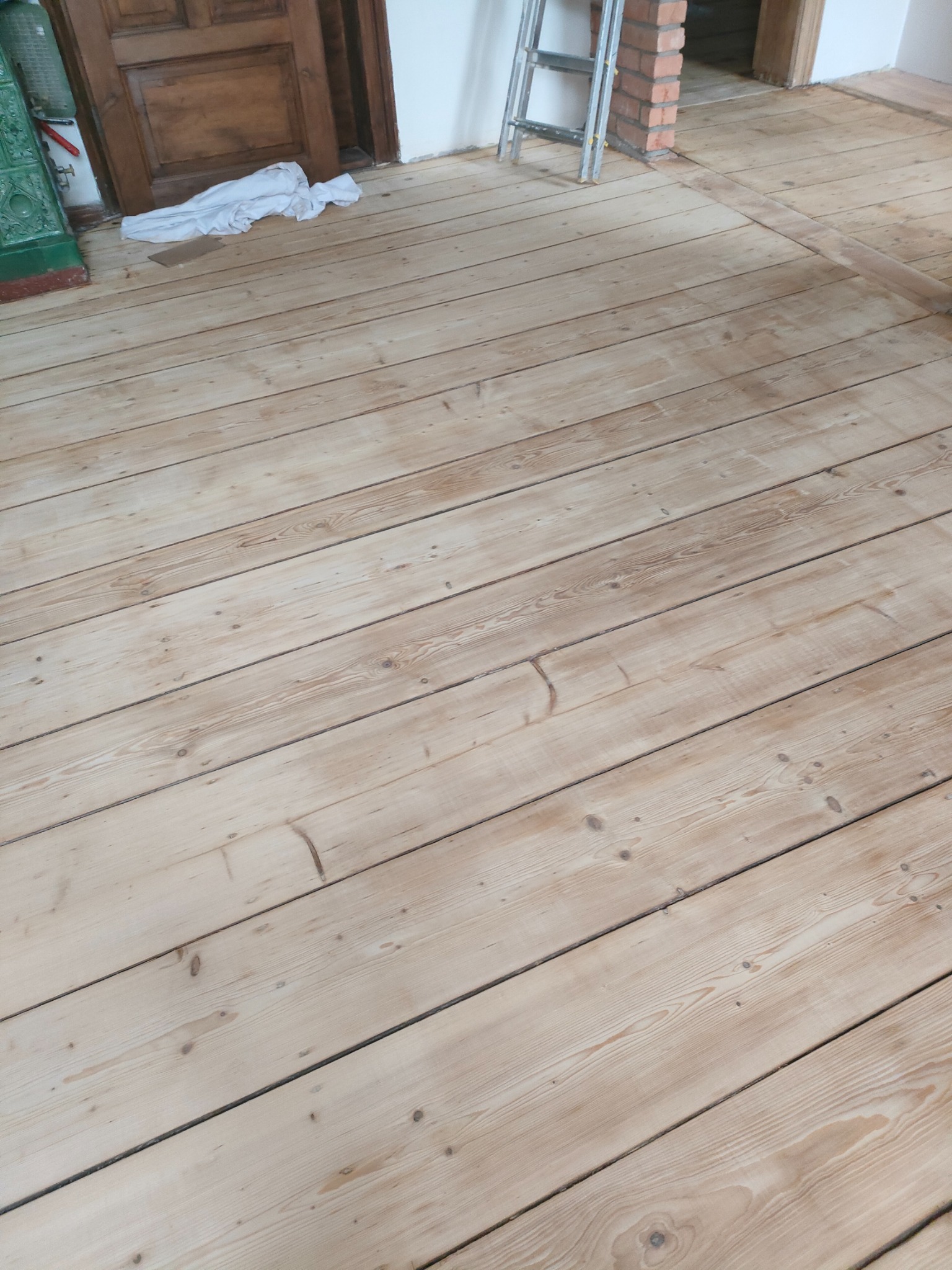 Jasna, odnowiona podłoga z desek po cyklinowaniu, widoczne ślady po szpachlowaniu, drabina oparta o ścianę w tle, drzwi z ciemnego drewna po lewej stronie.