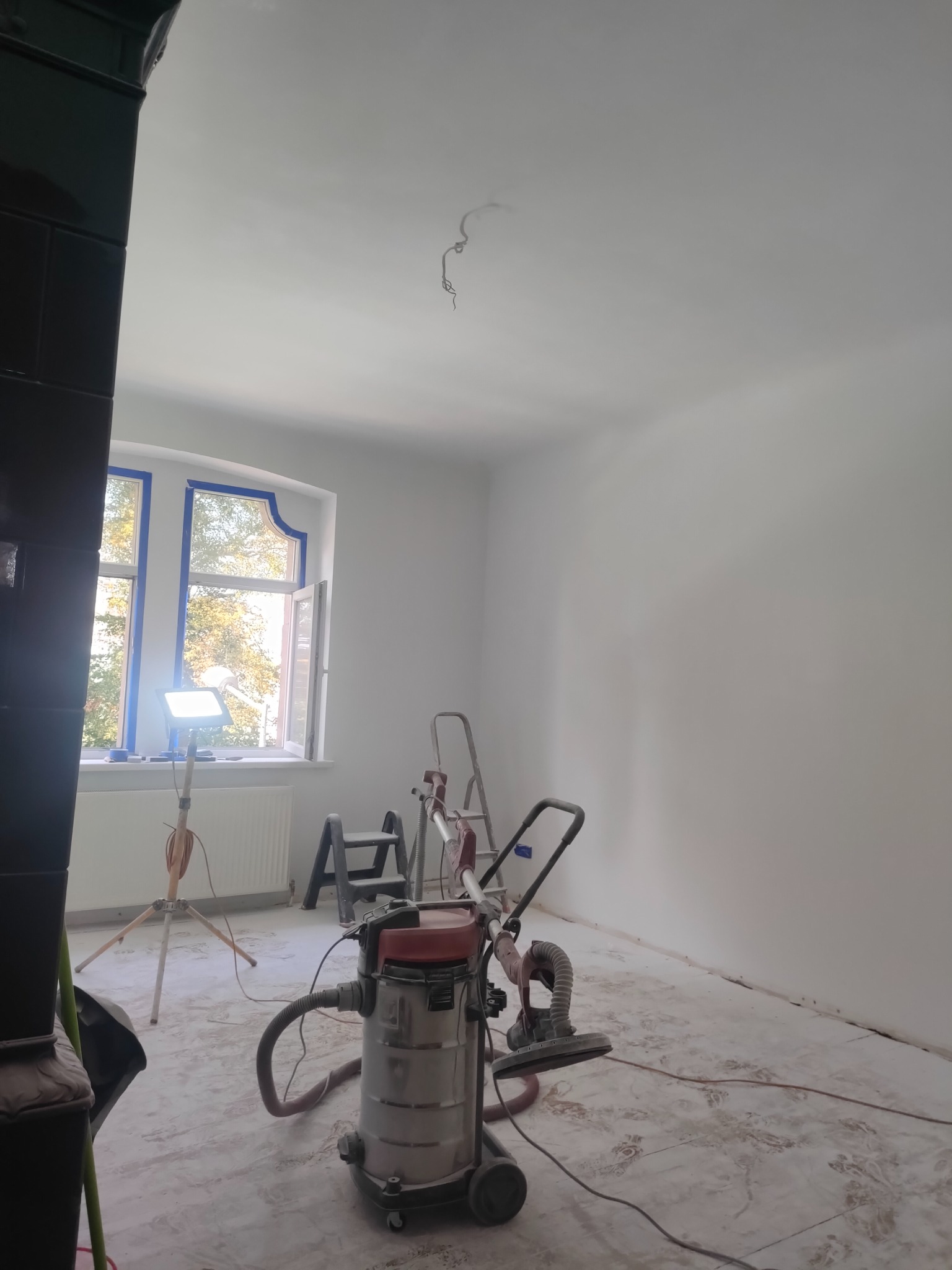 Białe ściany po szpachlowaniu w pokoju z oknem zabezpieczonym taśmą malarską. Widoczny sprzęt do szlifowania gładzi i drabiny.