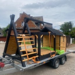 Go - Jumba Jakub Nowakowski - Transport domku zabaw dla dzieci na przyczepie, zabezpieczonego pomarańczowymi pasami. W tle ceglany budynek i pochmurne niebo.