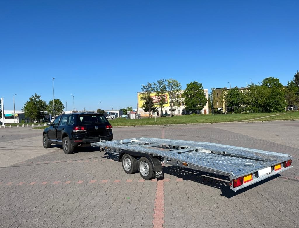 Czarny SUV z podpiętą, dwuosiową, aluminiową przyczepą na parkingu w słoneczny dzień. Widok z tyłu, z budynkami w tle.