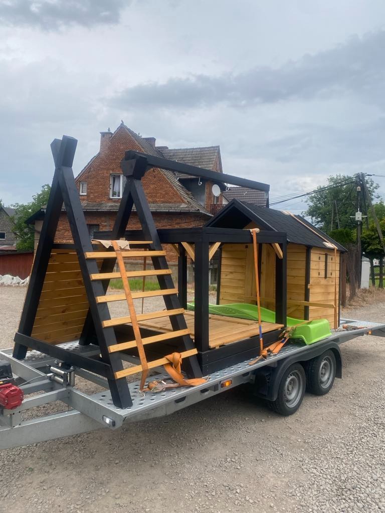 Transport domku zabaw dla dzieci na przyczepie, zabezpieczonego pomarańczowymi pasami. W tle ceglany budynek i pochmurne niebo.