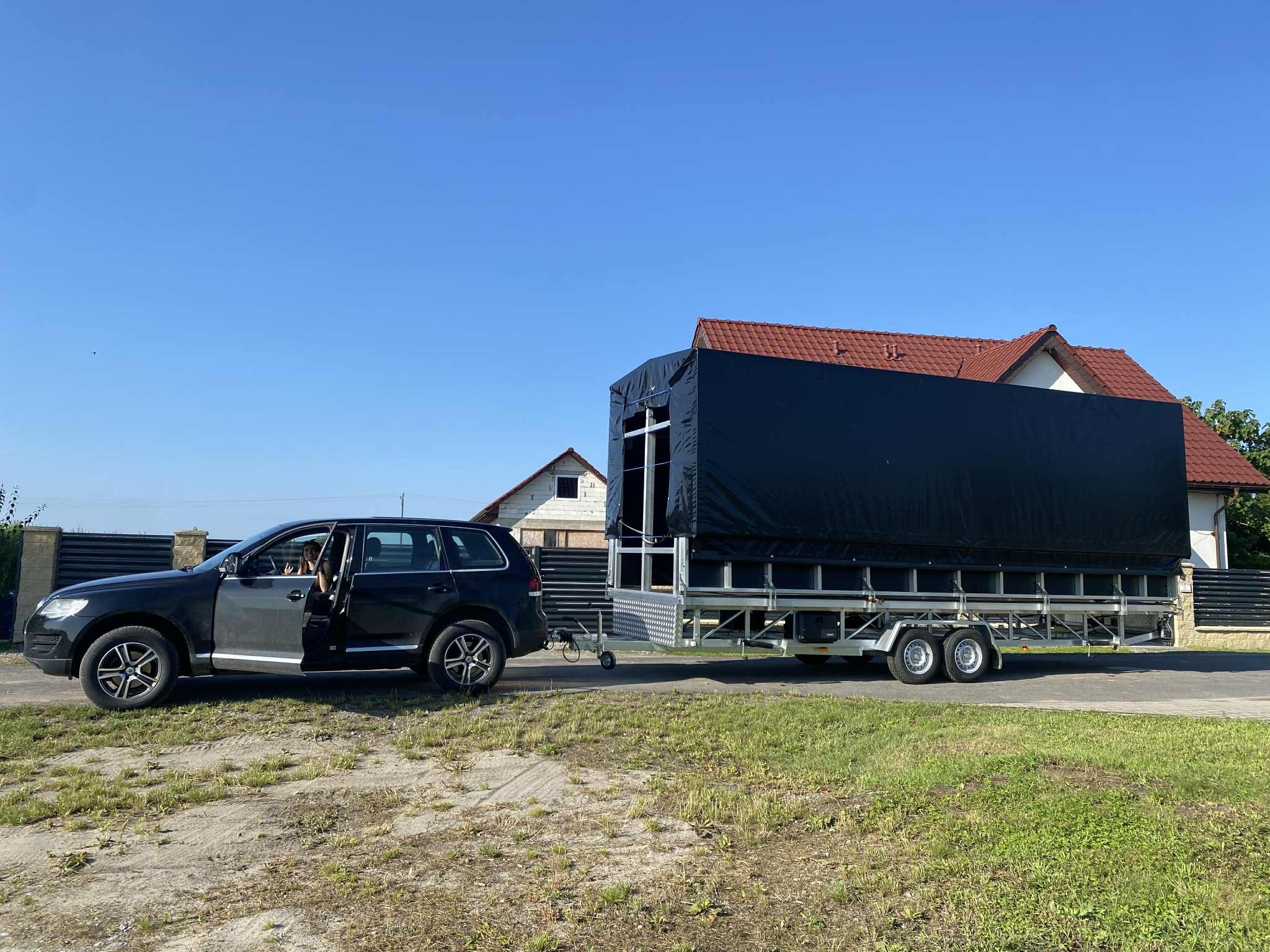 Czarny SUV z otwartymi drzwiami ciągnie dużą, zakrytą przyczepę na dwuosiowym podwoziu, na tle domów i błękitnego nieba. Widok z boku, w dzień.