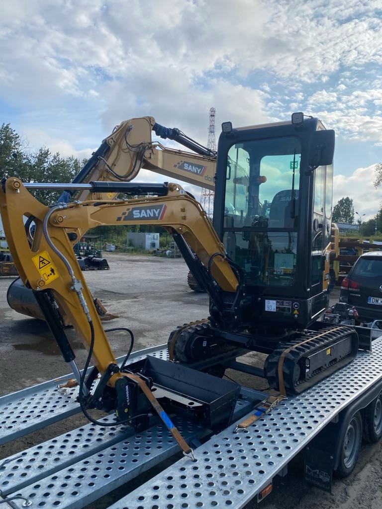 Żółta mini koparka SANY zabezpieczona pasami na perforowanej platformie lawety w Oławie. Widoczne niebo z chmurami i inne maszyny w tle.