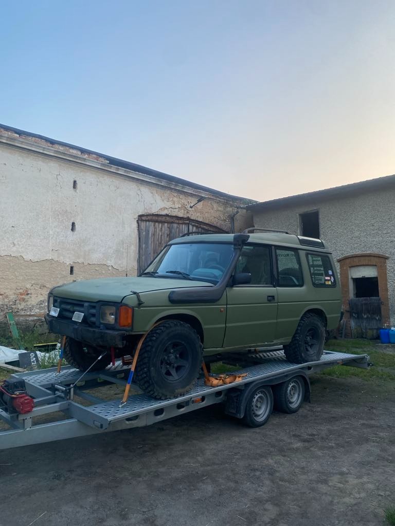 Laweta przewozi terenowy samochód Land Rover Discovery w kolorze khaki na tle wiejskiego krajobrazu z budynkami gospodarczymi.