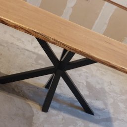 Adrian Malinowski - Stół z blatem z jasnego drewna o nieregularnych krawędziach, wsparty na czarnej, metalowej podstawie w kształcie gwiazdy. Surowe wykończenie wnętrza w tle.