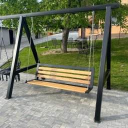 Adrian Malinowski - Metalowa huśtawka ogrodowa z drewnianym siedziskiem i dodatkowym wiszącym fotelem, na tle zielonego trawnika i kostki brukowej. Konstrukcja w kolorze czarnym.