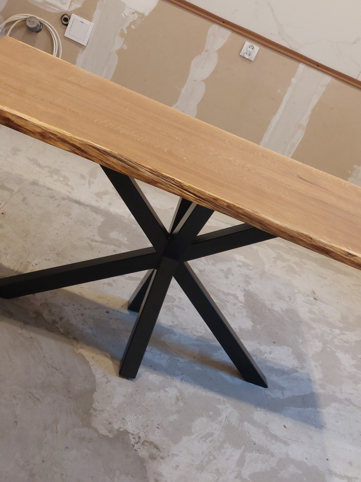 Stół z blatem z jasnego drewna o nieregularnych krawędziach, wsparty na czarnej, metalowej podstawie w kształcie gwiazdy. Surowe wykończenie wnętrza w tle.