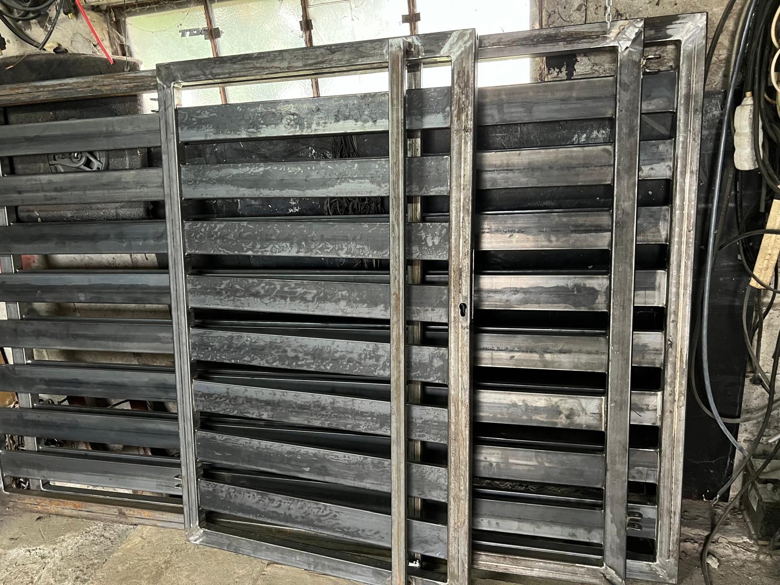 Surowe, metalowe elementy bramy wjazdowej z poziomymi lamelami, w trakcie produkcji. Widoczne spawy i niedoskonałości obróbki, surowy stan materiału.