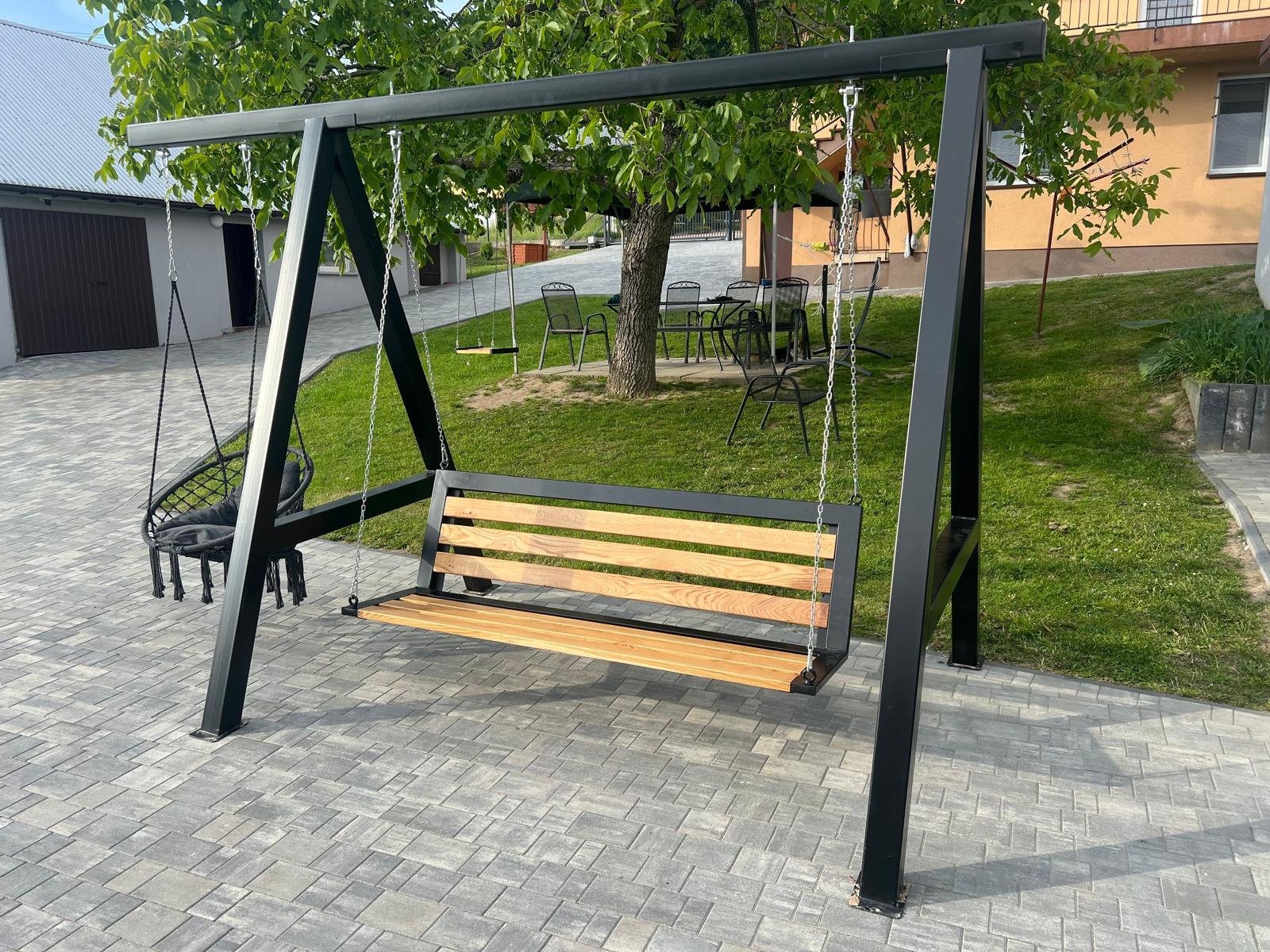 Metalowa huśtawka ogrodowa z drewnianym siedziskiem i dodatkowym wiszącym fotelem, na tle zielonego trawnika i kostki brukowej. Konstrukcja w kolorze czarnym.