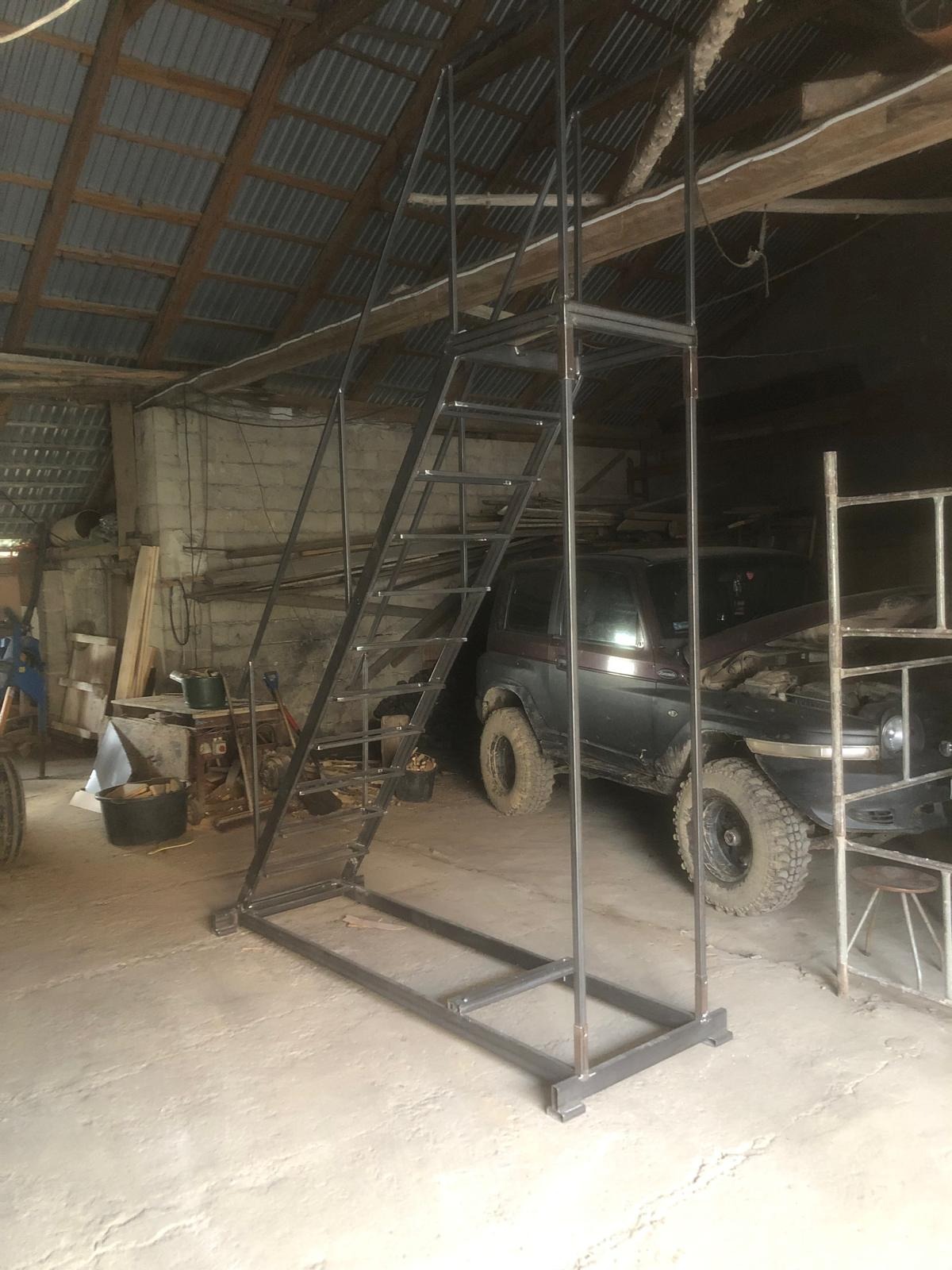 Surowa, metalowa konstrukcja schodów z platformą, spawana w warsztacie. W tle widoczny samochód terenowy i narzędzia.