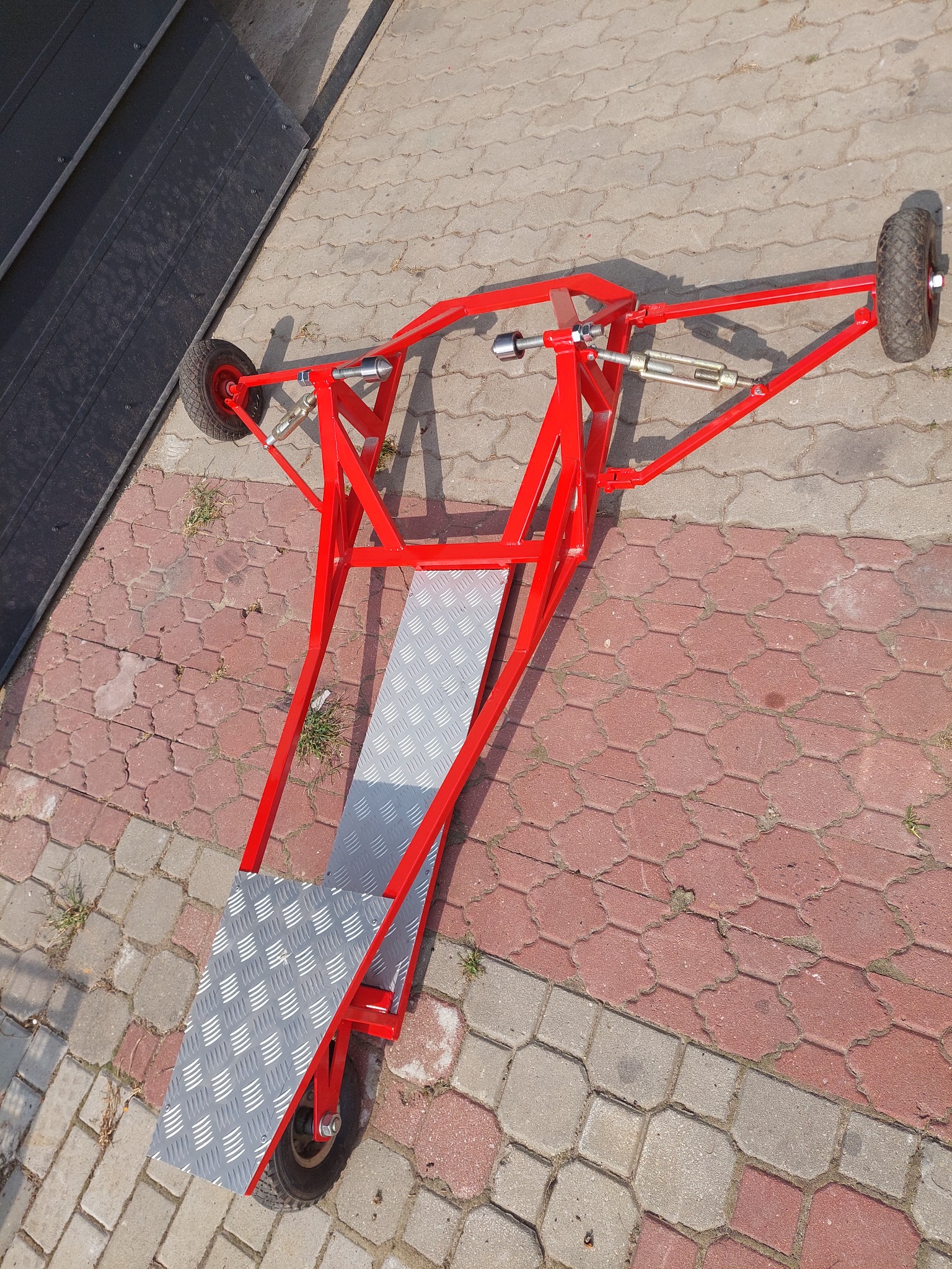 Czerwona, metalowa konstrukcja na trzech kołach z platformą z blachy ryflowanej, leżąca na kostce brukowej. Widoczne elementy śrubowe i regulacyjne.