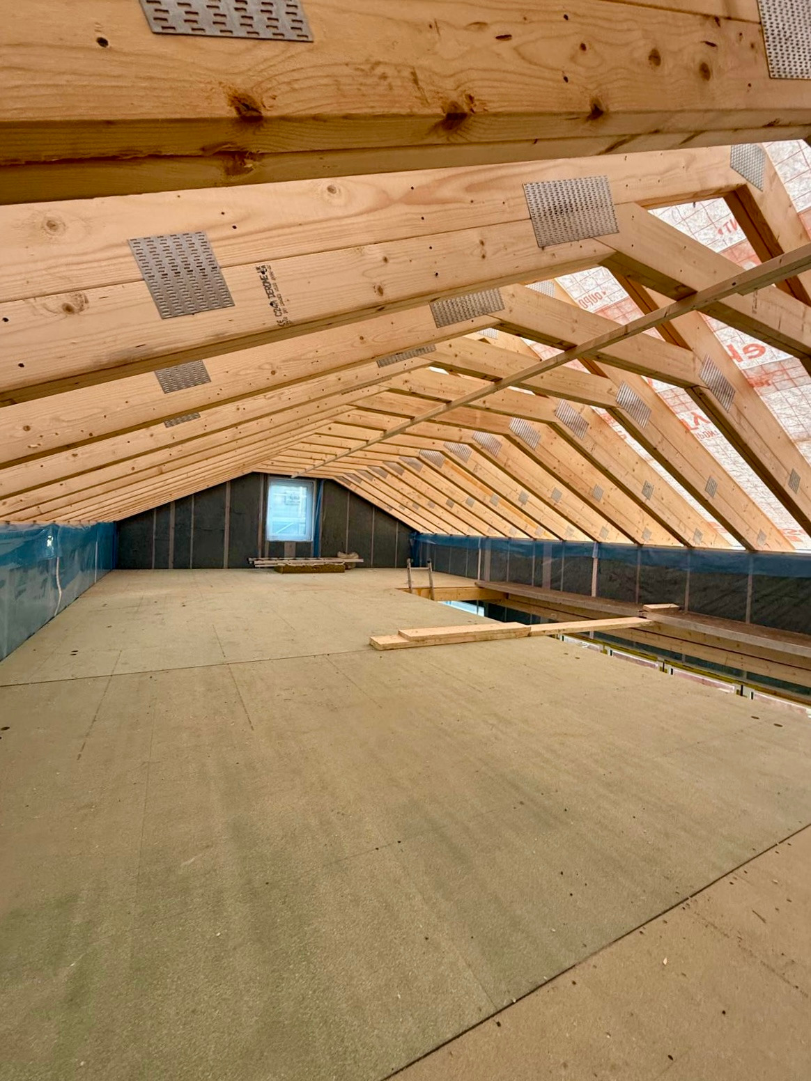 Wnętrze poddasza z drewnianą konstrukcją dachu, ocieplone wełną mineralną i zabezpieczone folią. Widoczne metalowe łączniki belek i okno w szczycie.