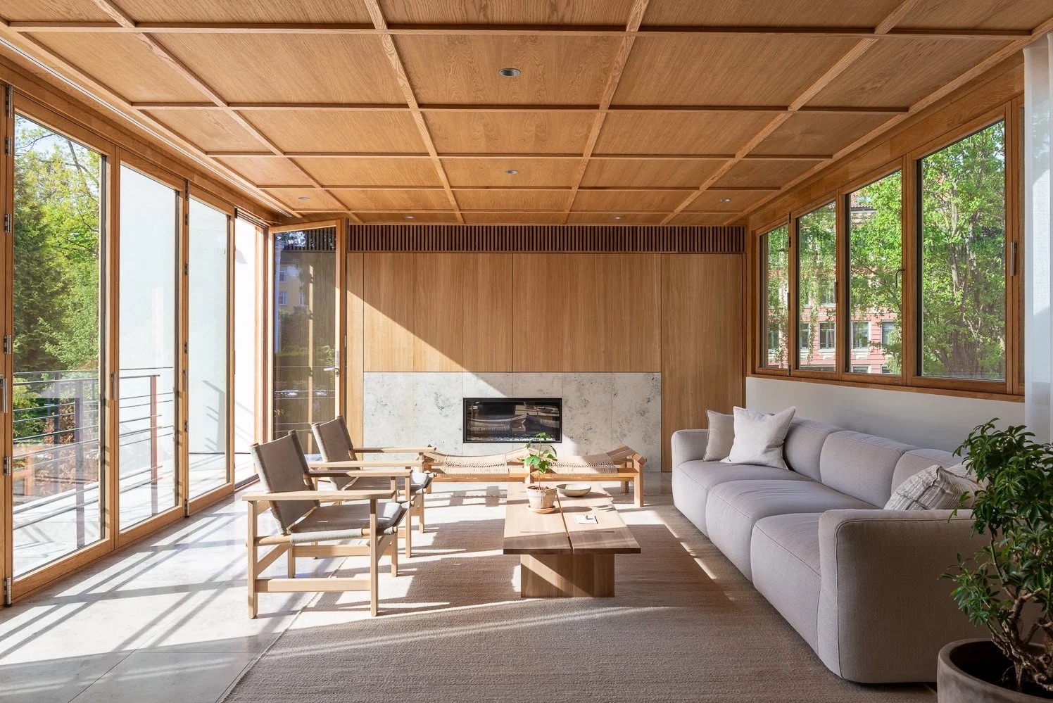 Jasne wnętrze z drewnianym sufitem kasetonowym i dużymi oknami. Minimalistyczny salon z kominkiem i meblami w naturalnych odcieniach. Wpadające promienie słoneczne.