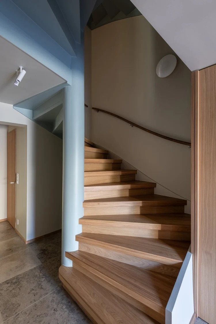 Drewniane schody spiralne z kolumną w kolorze pastelowego błękitu, oświetlone światłem dziennym. Wykończenie wnętrz w nowoczesnym stylu, Poznań.