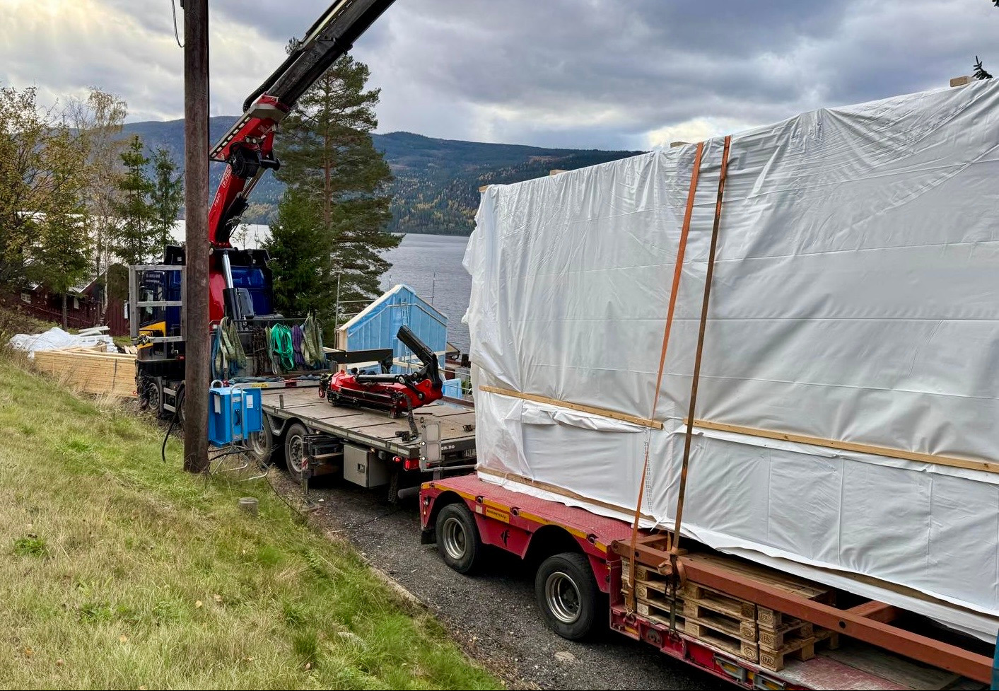 Transport modułu domu szkieletowego na lawecie z użyciem dźwigu, zabezpieczonego folią, na tle jeziora i zieleni. Widoczne elementy konstrukcyjne i palety.