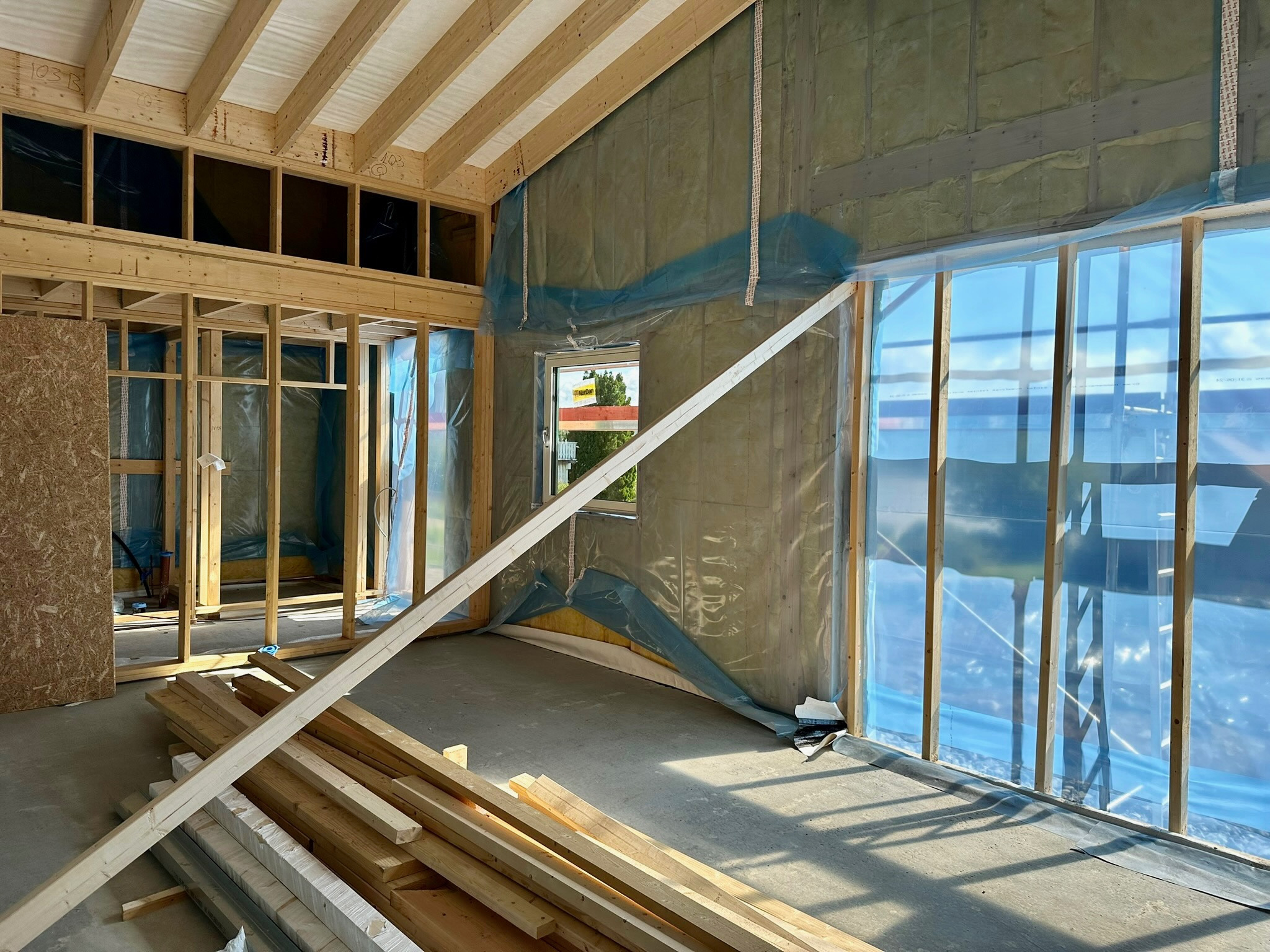 Wnętrze domu w trakcie budowy: drewniany szkielet, ocieplone ściany zabezpieczone folią, przygotowane deski. Widoczne elementy konstrukcyjne i materiały budowlane.