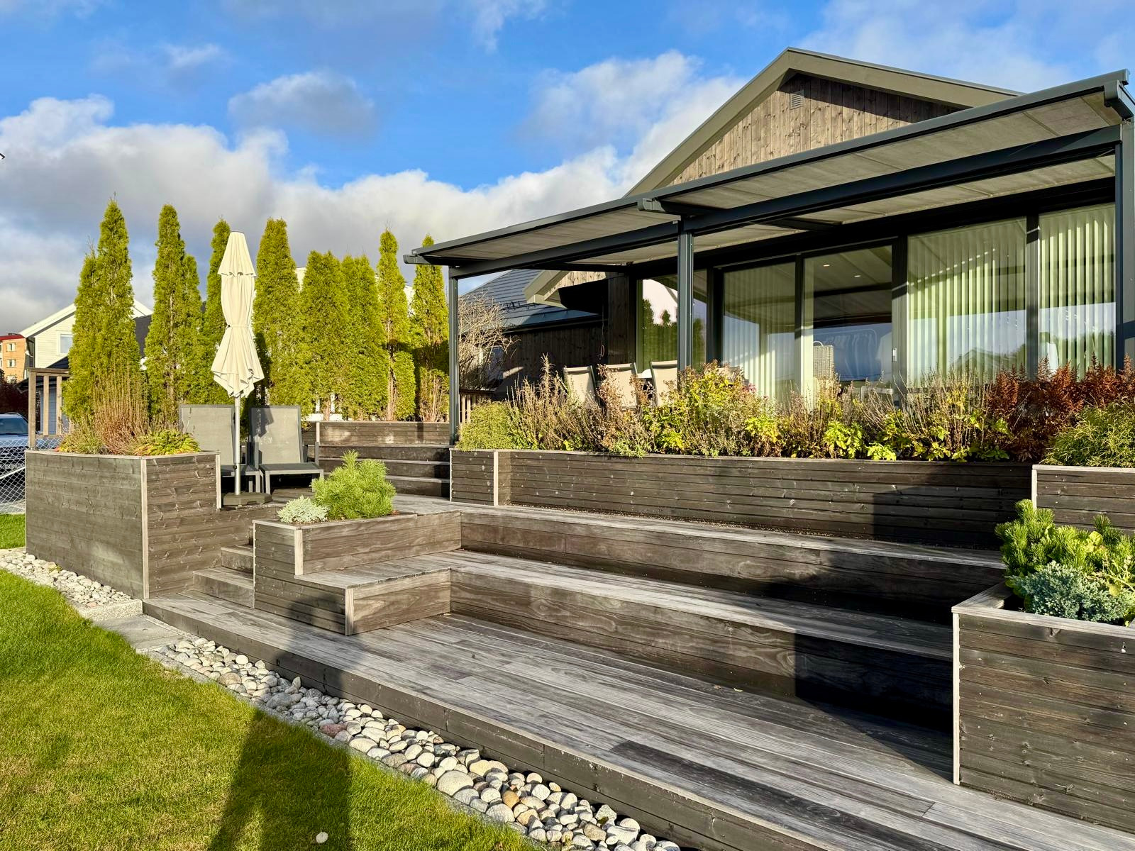 Drewniany taras z roślinami w donicach, schody z desek, kamienne obrzeże, nowoczesny dom z dużymi oknami i zadaszeniem, zielony trawnik, błękitne niebo z chmurami.