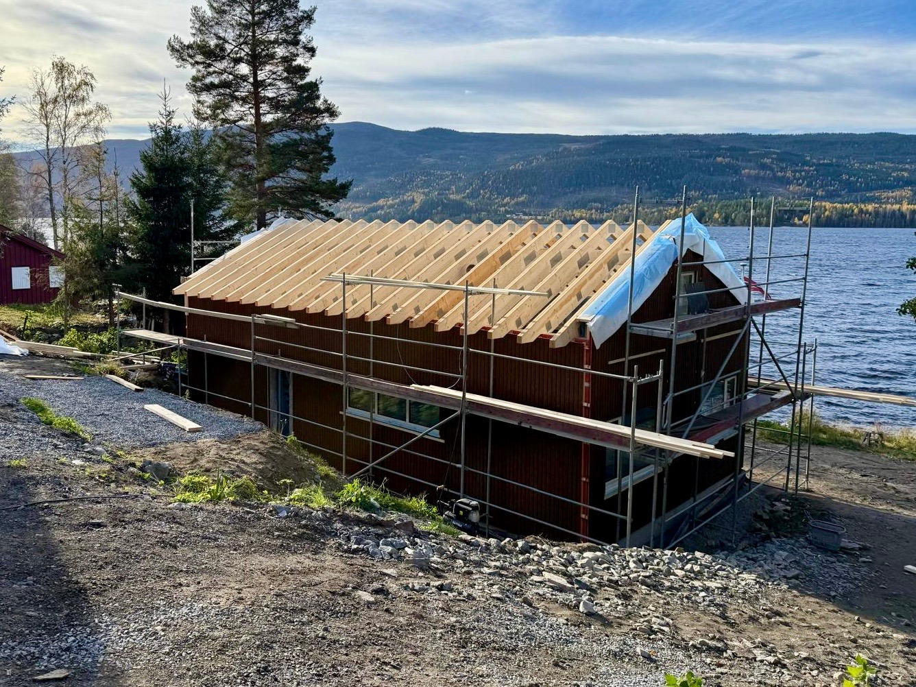 Budowa domu w stylu skandynawskim z drewnianą konstrukcją dachu, zabezpieczoną folią, na tle jeziora i gór, widoczne rusztowania.