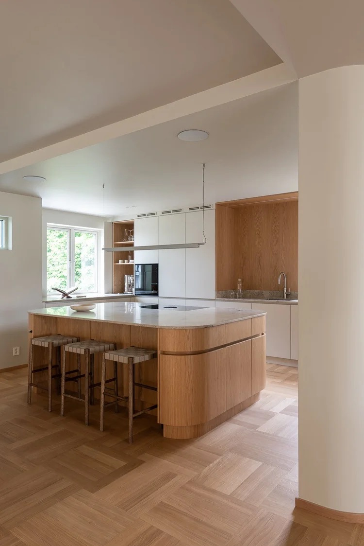 Jasna kuchnia z wyspą o zaokrąglonych frontach z drewna, parkiet w jodełkę i minimalistyczne oświetlenie. Nowoczesny design z naturalnymi materiałami.