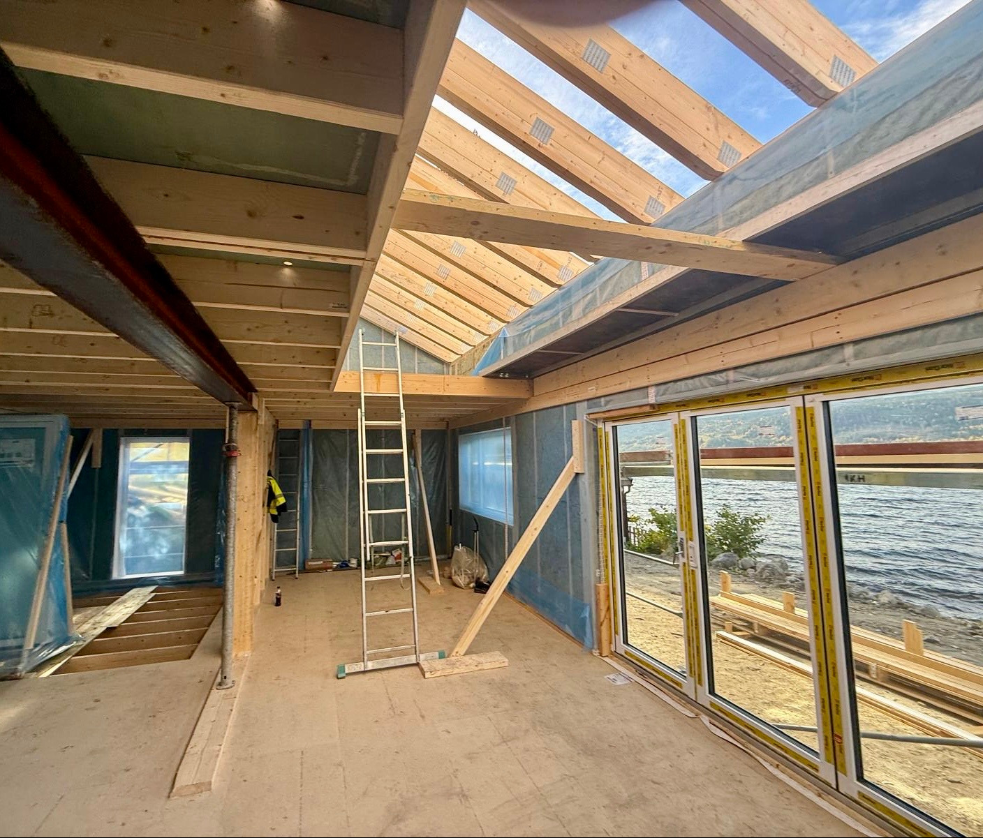 Wnętrze domu szkieletowego w trakcie budowy z widokiem na jezioro przez duże okna. Widoczne drewniane elementy konstrukcyjne i drabina.