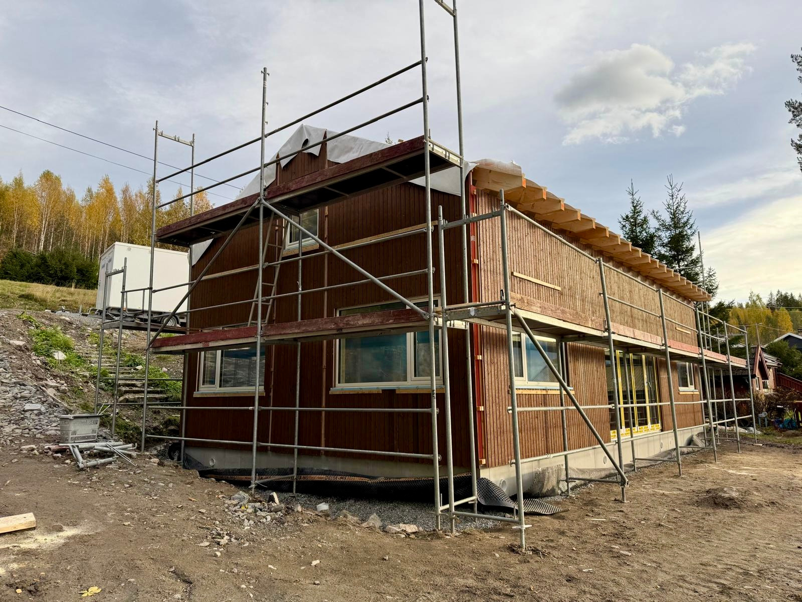 Brązowy dom w trakcie budowy, otoczony rusztowaniami. Widoczne drewniane deski elewacyjne i okna. W tle drzewa i niebo z chmurami. Etap prac wykończeniowych.