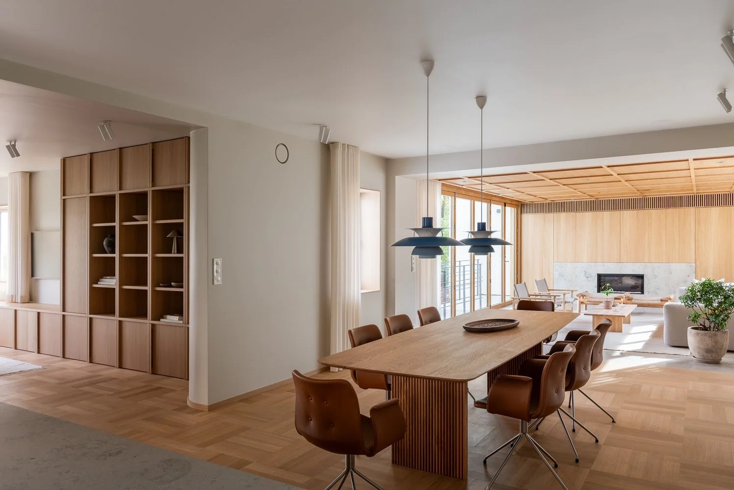 Jasne, przestronne wnętrze z drewnianą podłogą w jodełkę, dużym stołem jadalnym i zabudową meblową z drewna. Widoczne designerskie lampy i kominek.