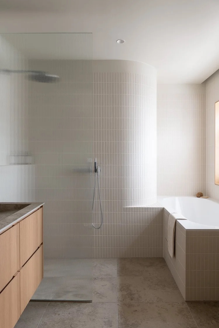 Minimalistyczna łazienka z kabiną prysznicową ze szkła ryflowanego, beżowymi płytkami ułożonymi pionowo i drewnianą szafką pod umywalką. Widoczna wanna i szara podłoga.