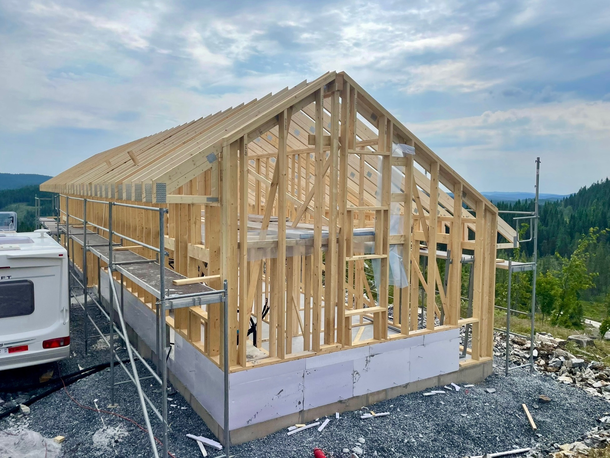 Surowy szkielet drewnianego domu w trakcie budowy, z widocznymi rusztowaniami i zielonym tłem lasu. Elementy konstrukcyjne z jasnego drewna.