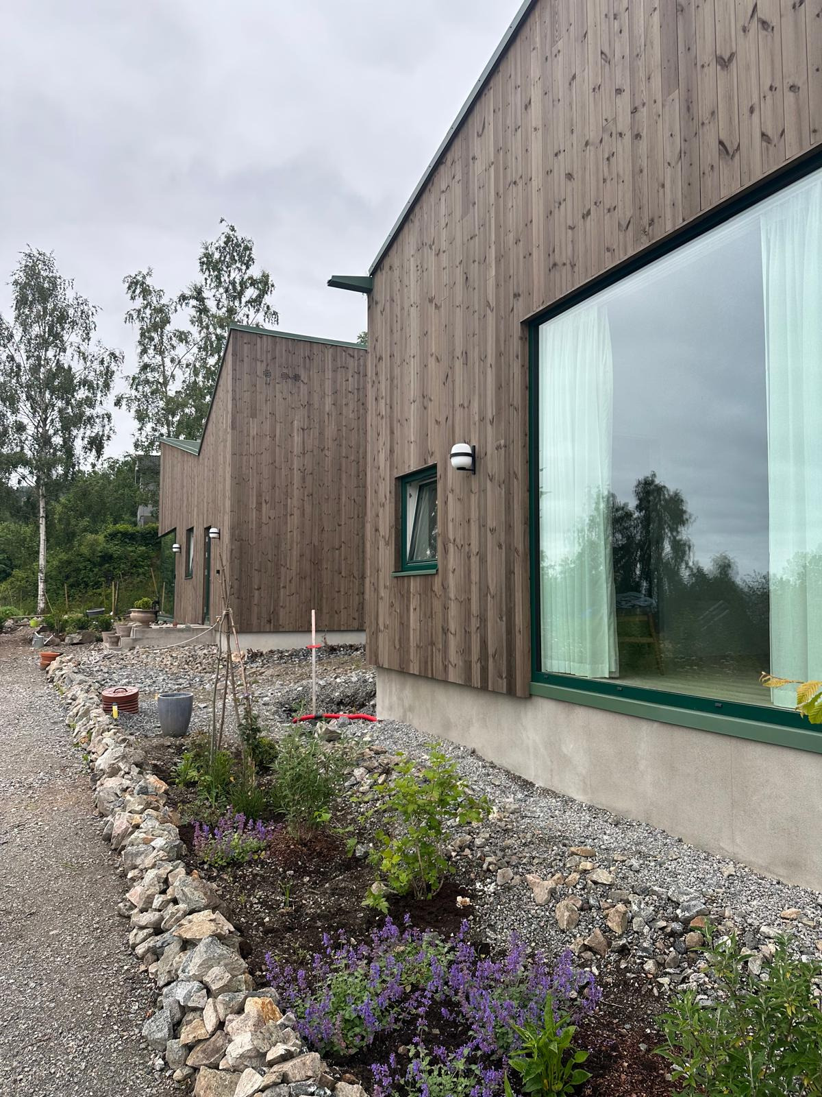 Dwa drewniane domy z dużymi oknami i zielonymi ramami, otoczone kamieniami i roślinami. Widoczny fragment ścieżki i drzewa w tle. Ujęcie z niskiej perspektywy.
